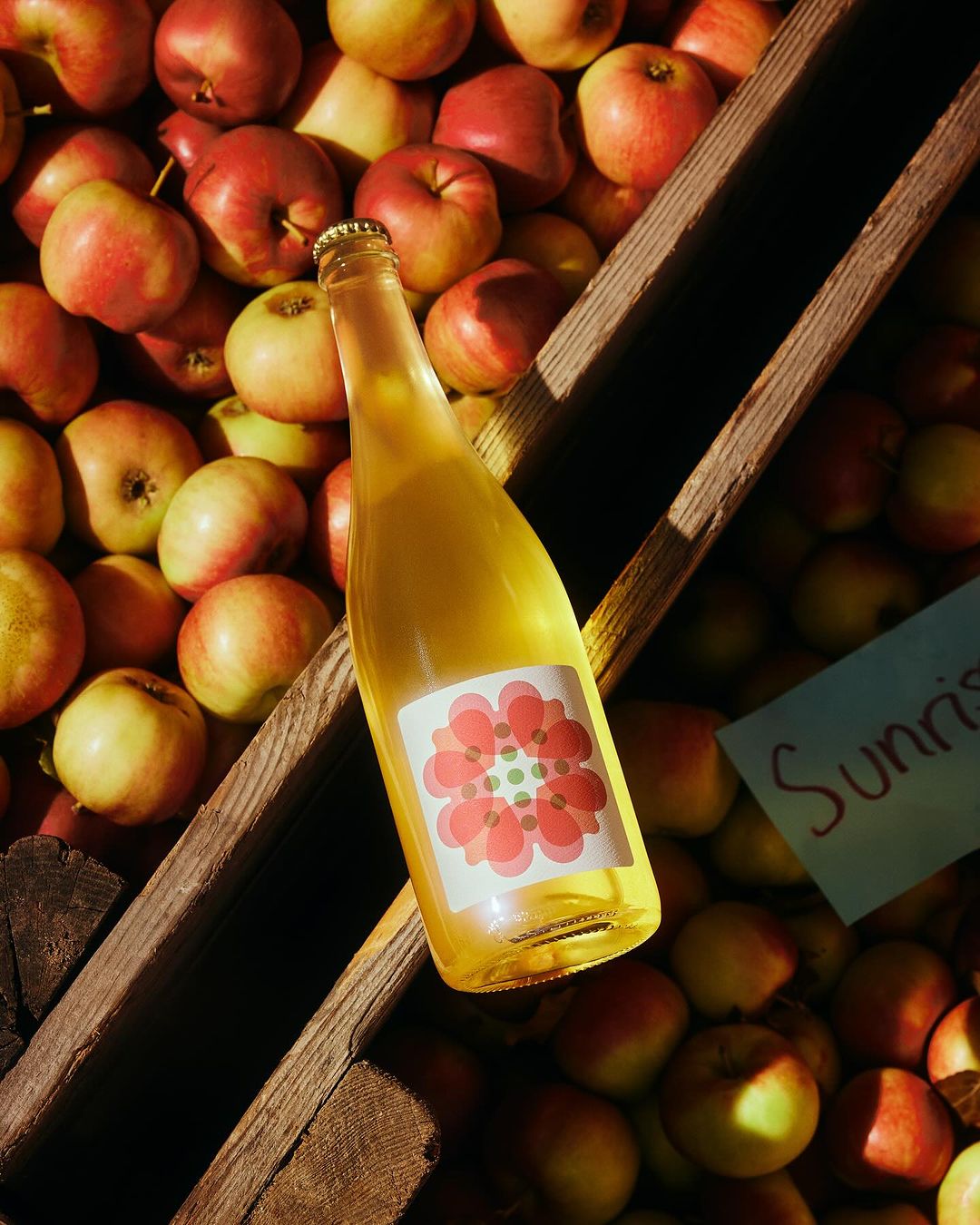 Bottle of apple cider displayed with fresh apples in a wooden container, bathed in soft, directional light.