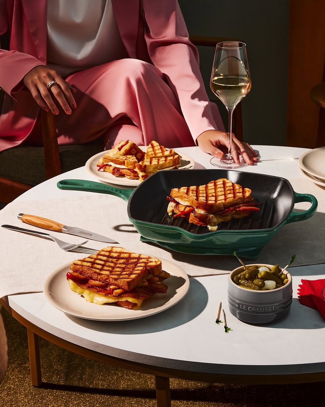 Elevated shot of grilled cheese sandwiches, pickles, and white wine on a round table with a woman in a pink suit.