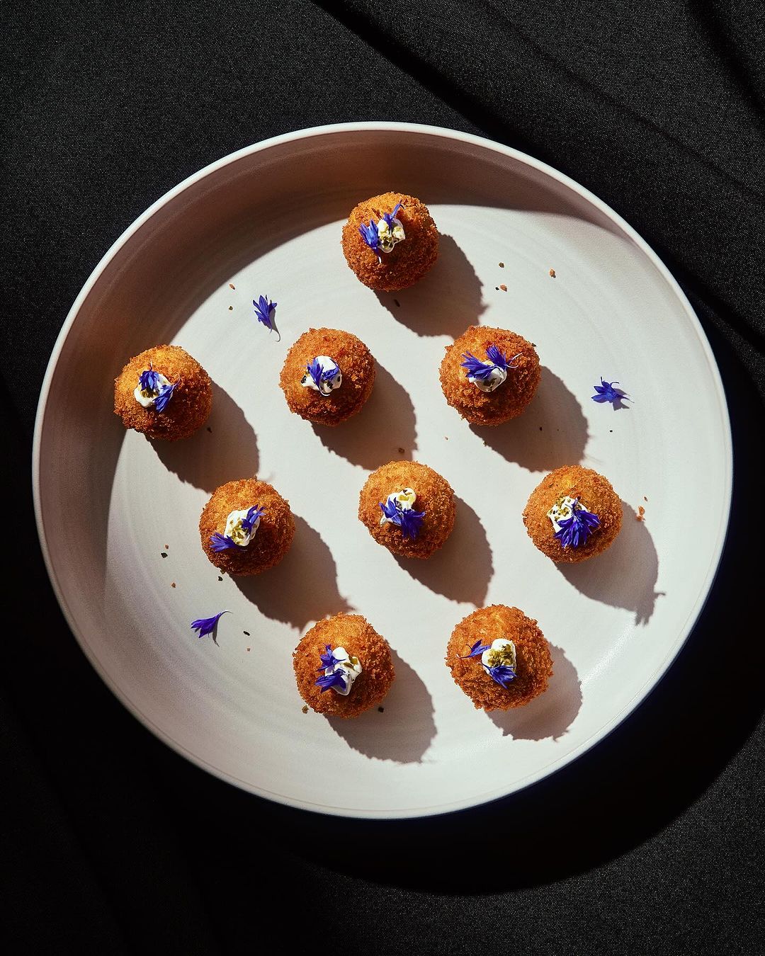 Overhead shot of fried arancini balls decorated with blue petals served on a white plate, placed against a dark fabric backdrop.