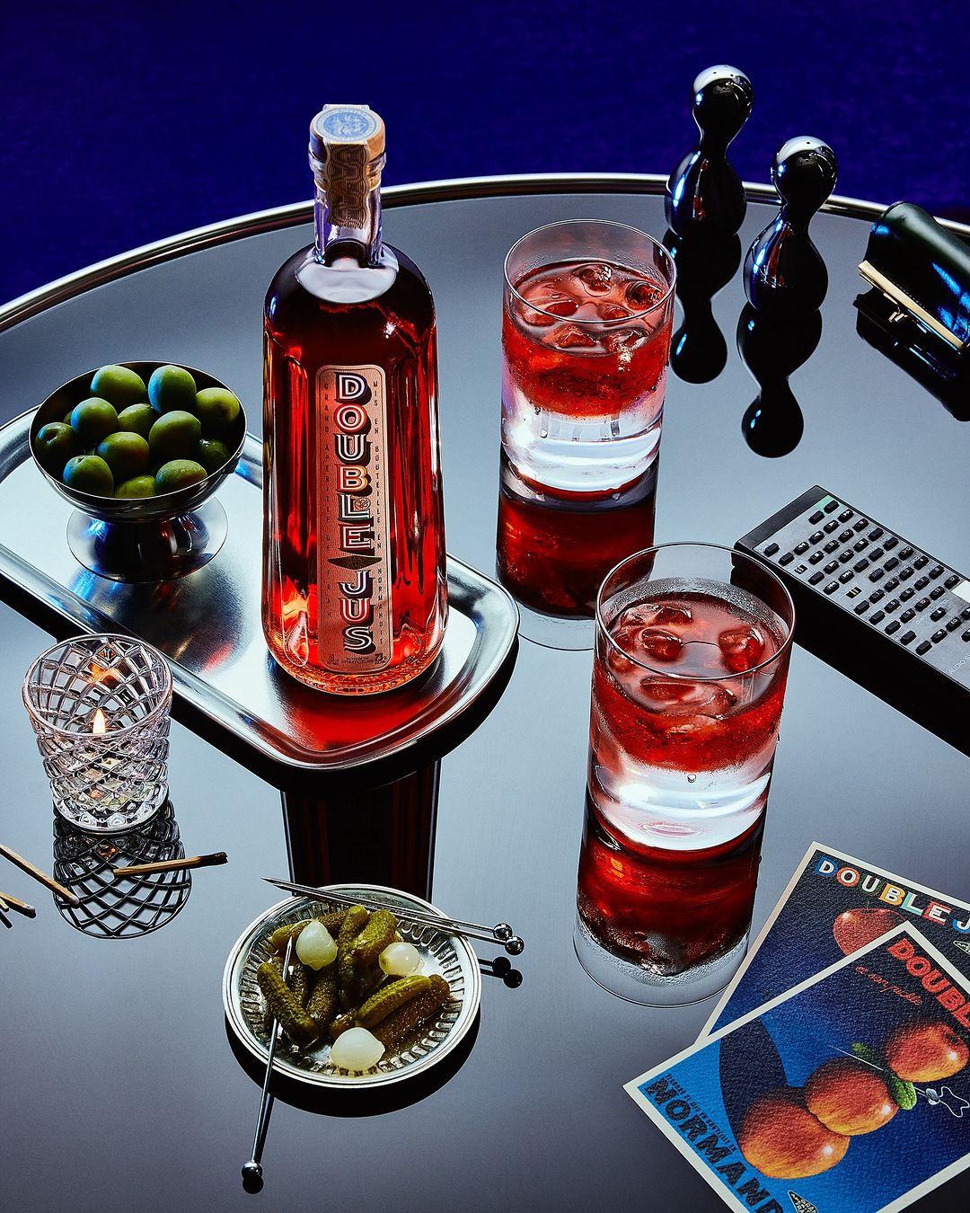Still life shot of a liquor bottle and two glasses with cocktails on a reflective table, garnished with olives, pickles, and a vintage poster.