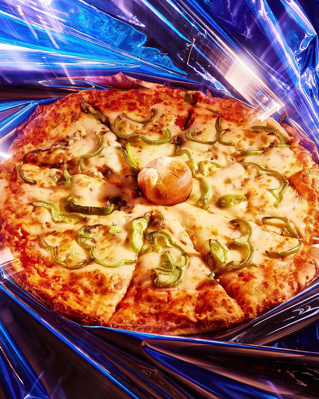 Overhead shot of a sliced pizza with green peppers and a bread roll in the center, resting on a metallic blue reflective surface.