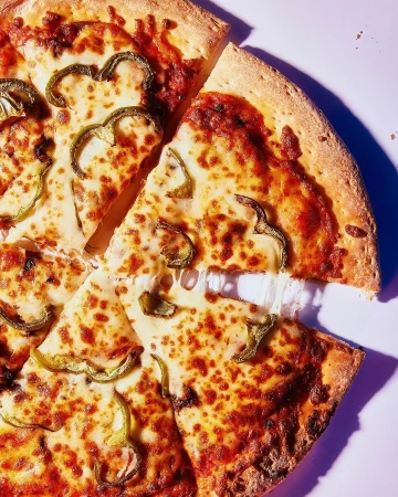 Overhead shot of a sliced pizza featuring melted cheese, tomato sauce, and green bell peppers on a light purple surface, food photography.