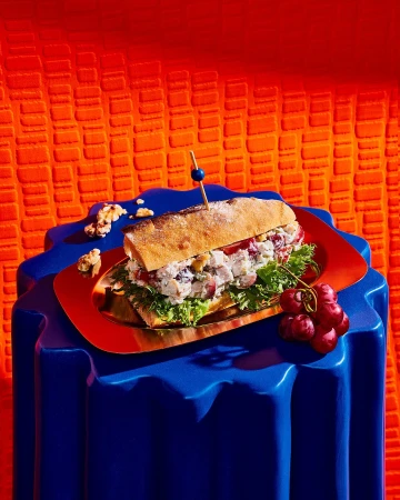 Overhead studio shot of a baguette chicken salad sandwich with walnuts and grapes on a copper tray with blue patterned table against orange backdrop.