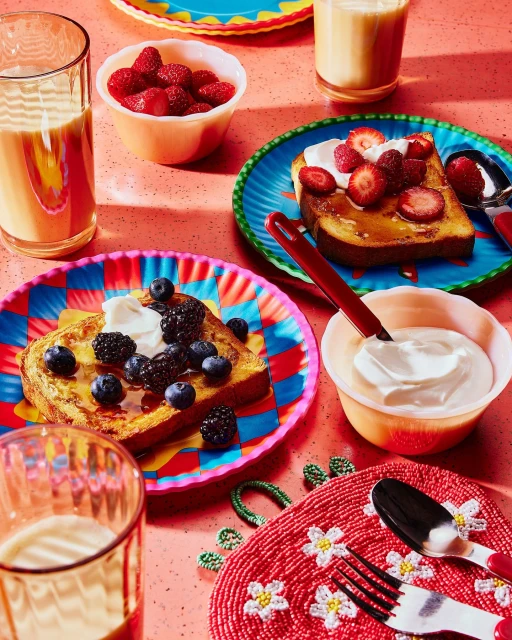 Overhead shot of french toast topped with fresh berries and cream, served on colorful plates with milk in a brightly lit indoor setting.