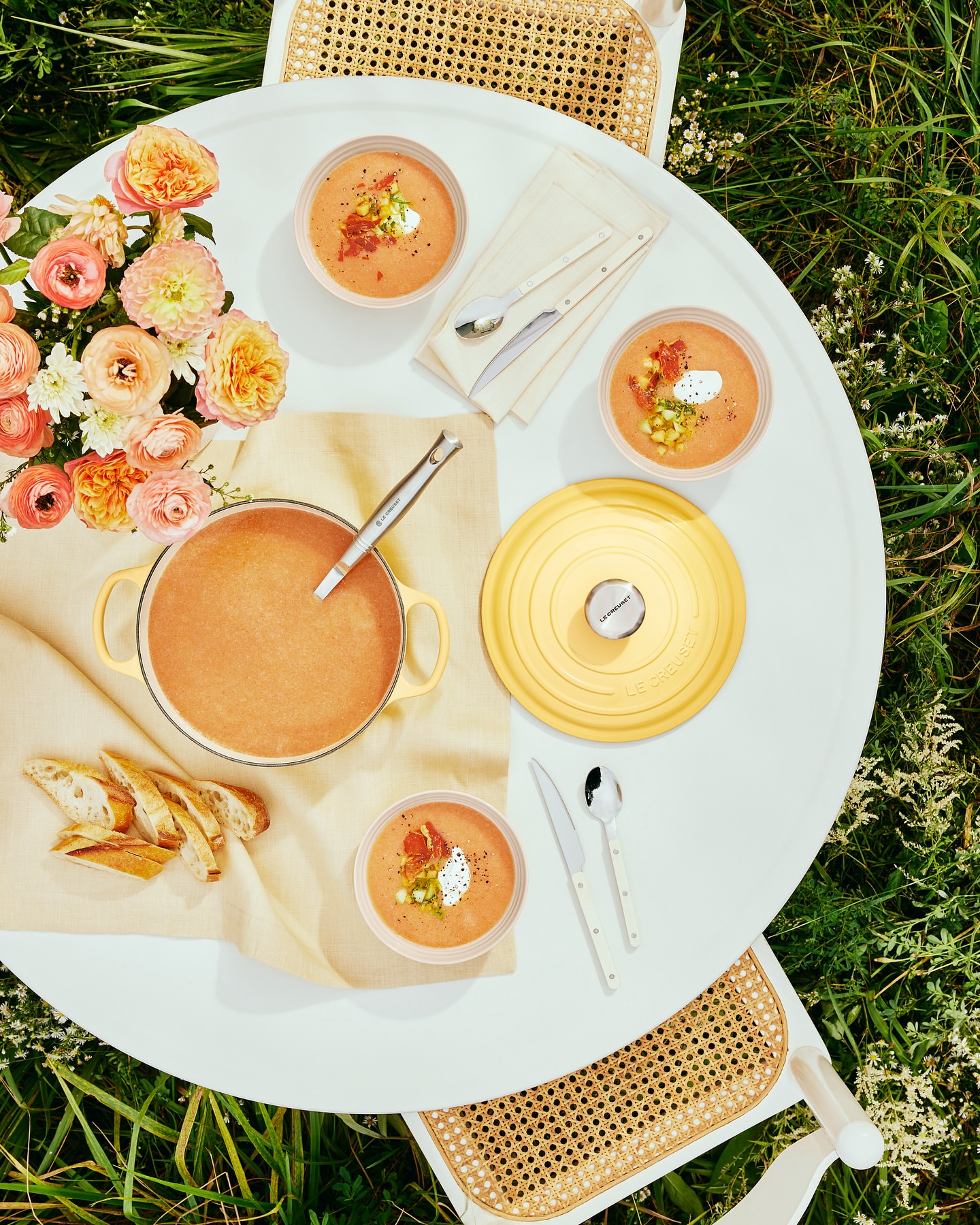 Overhead shot of a white table set with bowls of soup, a tureen, bread slices, silverware, and a bouquet of flowers on a grassy lawn.