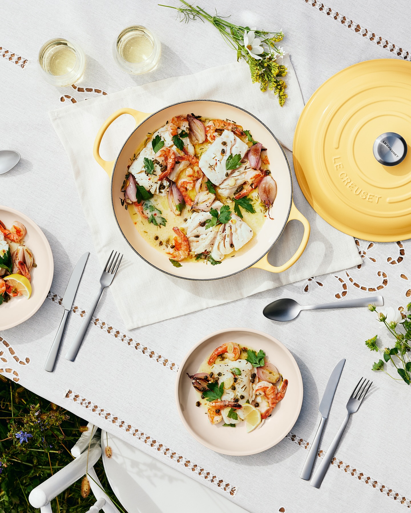Overhead shot of a table setting with a yellow cast iron pan and plated fish dishes, set on a white tablecloth with tableware and flowers.