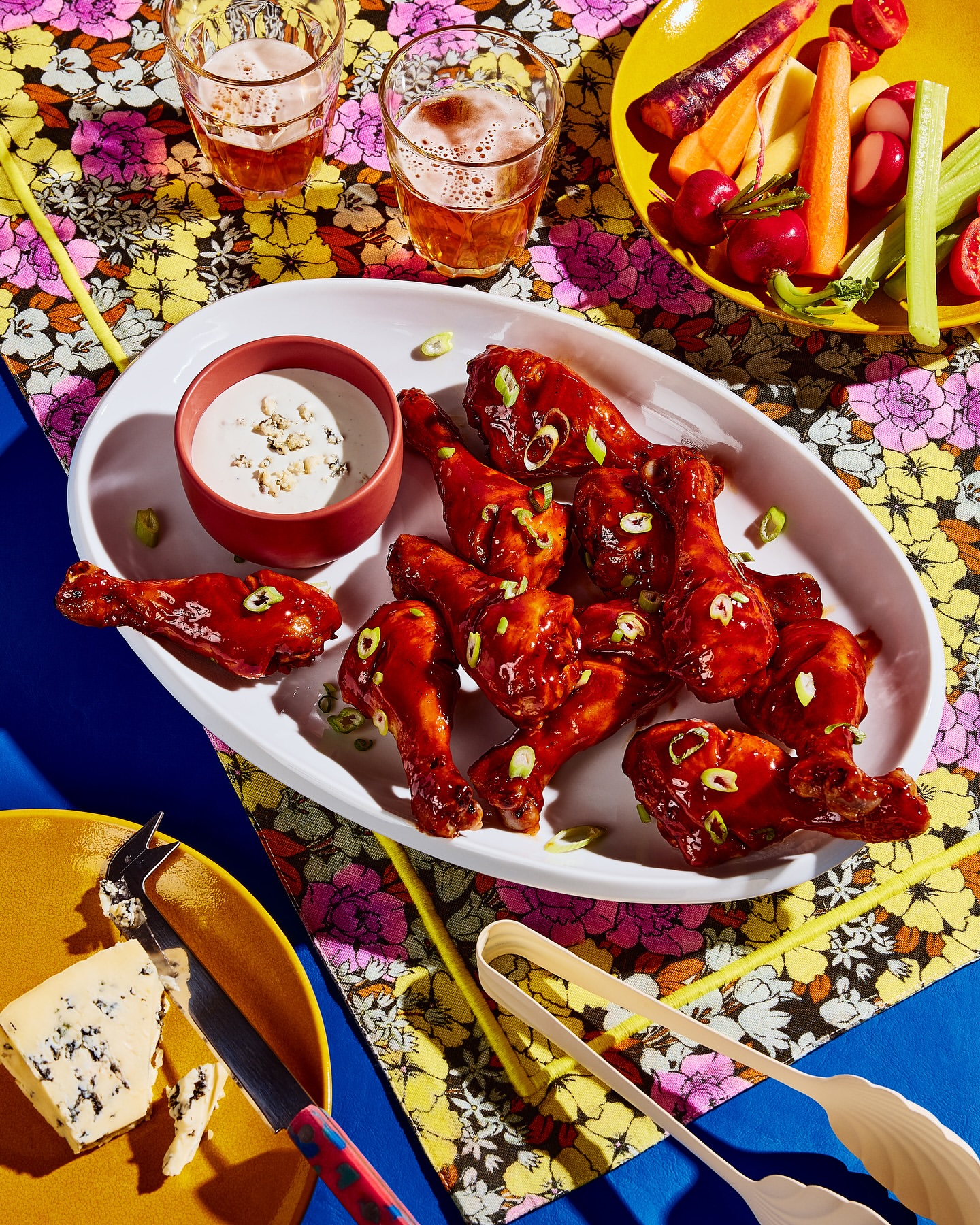 Overhead shot of glazed chicken drumsticks on a white platter with blue cheese dip, vegetable crudités, cheese, and drinks on a floral tablecloth.