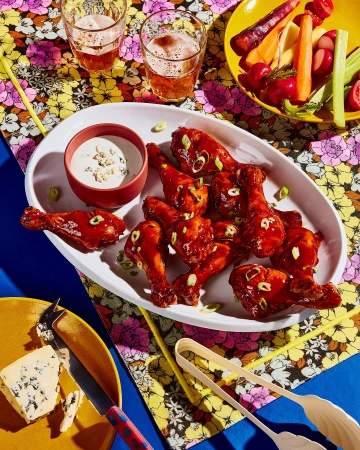 Overhead shot of glazed chicken drumsticks on a white platter with blue cheese dip, vegetable crudités, cheese, and drinks on a floral tablecloth.