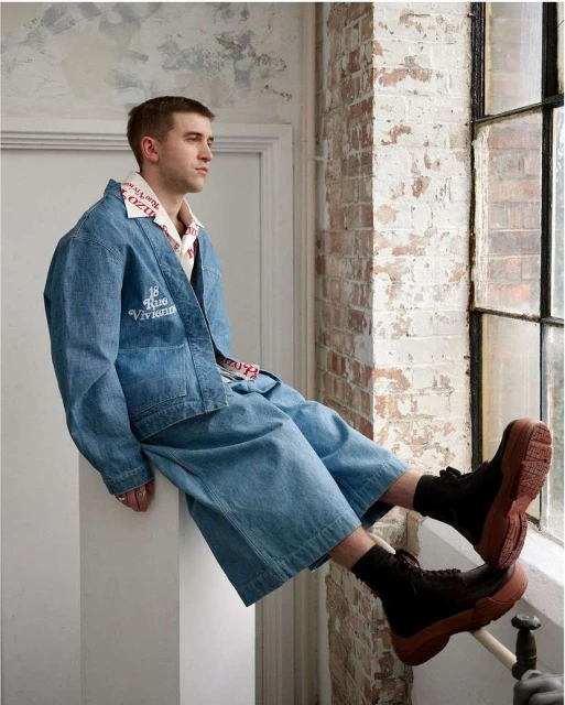 A young adult male sits by a window wearing an oversized denim jacket and wide-leg pants, paired with black socks and platform boots.