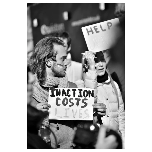 Monochrome shot of a group protesting, with a man in the foreground holding a sign reading 'INACTION COSTS LIVES.'