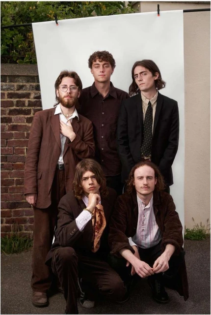 Five young adults in suits pose against white backdrop near a brick wall, three standing, two kneeling, in a medium outdoor shot.