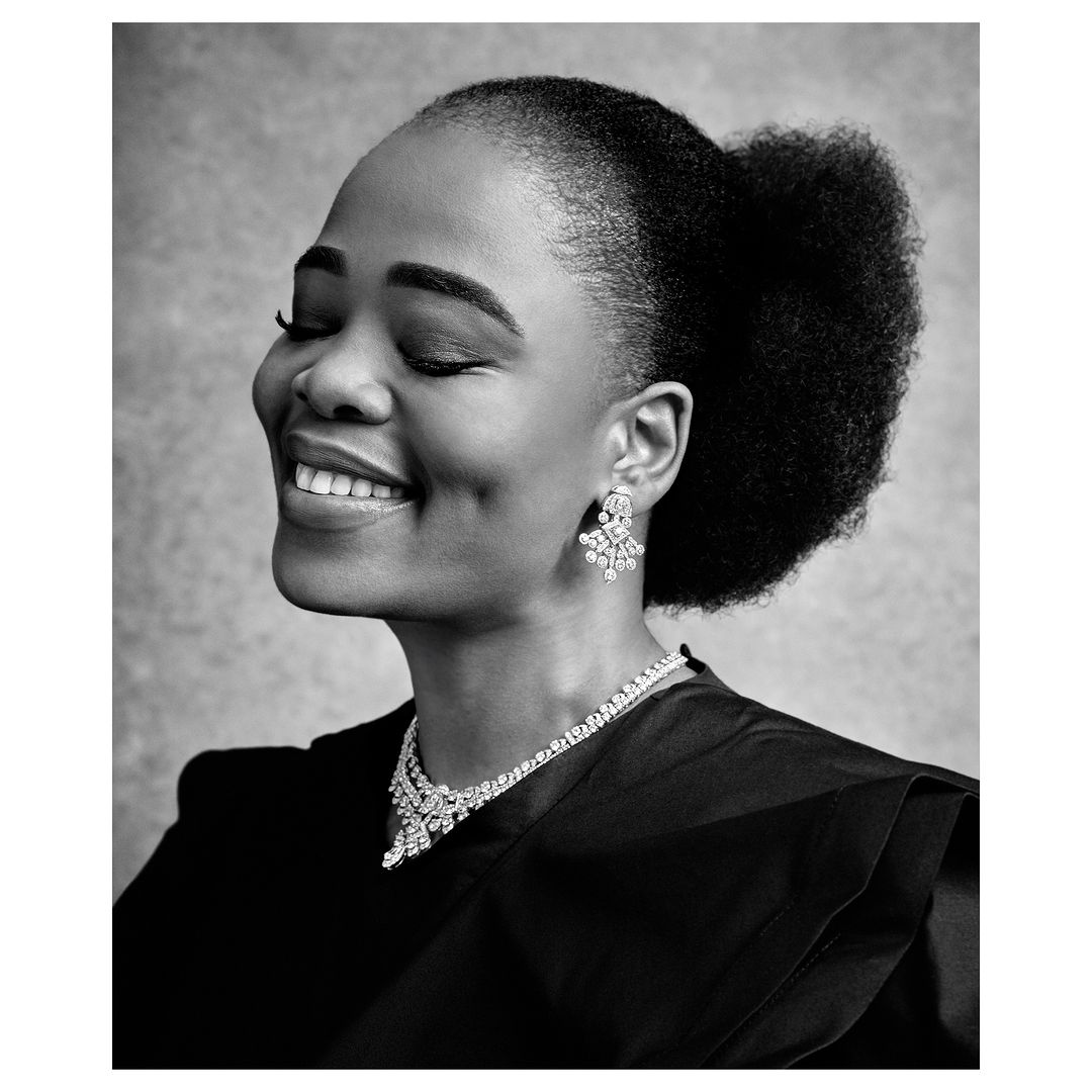 Black and white close-up portrait of a Black woman smiling with her eyes closed, wearing a necklace and earrings in a neutral setting.