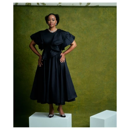 Black woman wearing a dark ruffle dress and heels standing on a white block against a textured green backdrop.