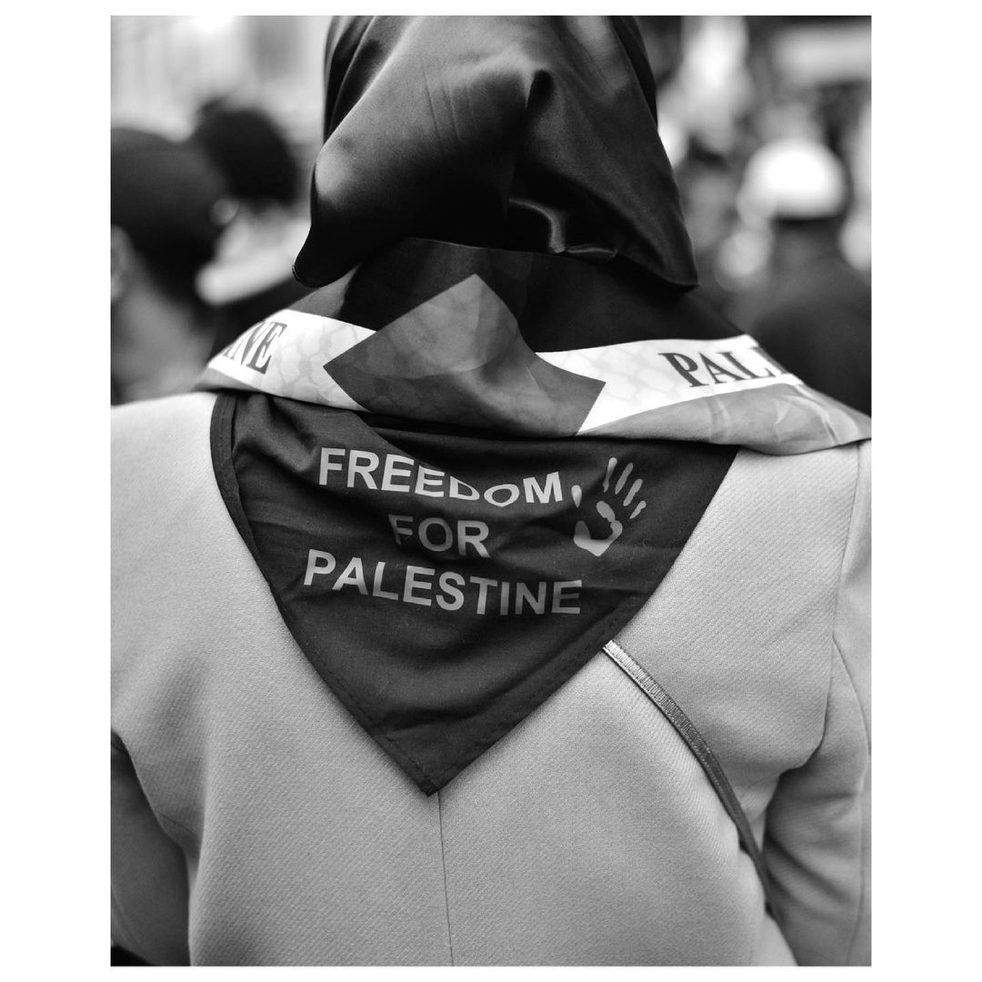 Person wearing a headscarf and a scarf with the words 'Freedom for Palestine' in black and white. Shoulder bag strap visible.