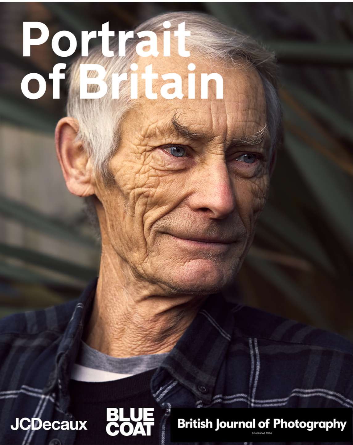 Close-up portrait of an elderly man with gray hair, wearing a blue and black plaid shirt, against a blurred outdoor background.