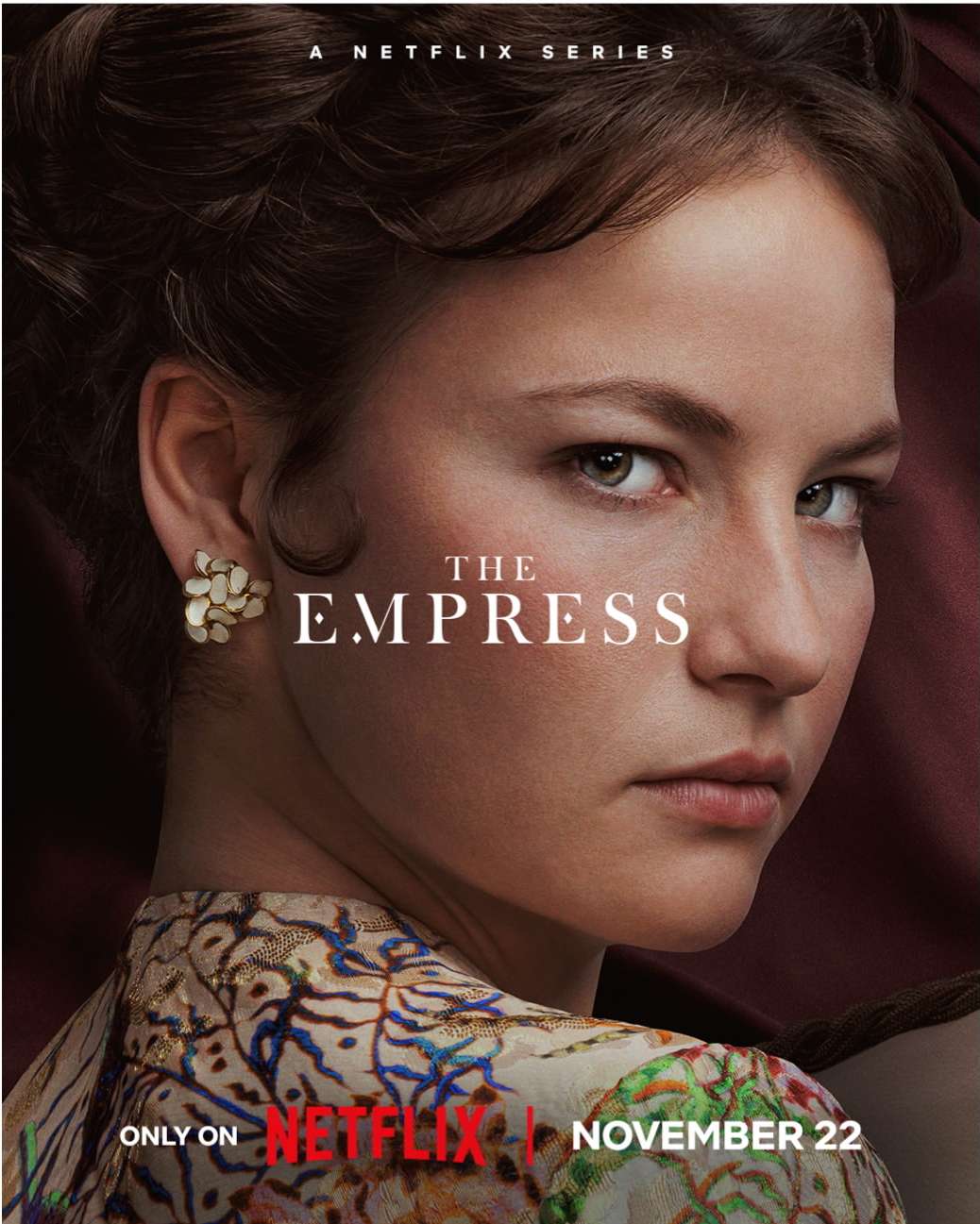 Close-up of a woman's face in The Empress Netflix series promo, she gazes intently forward, wearing a patterned top and ornate earrings.