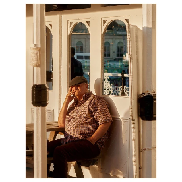 Mature man in cap and plaid shirt seated outdoors at wooden table. Natural light illuminates reflective window background.