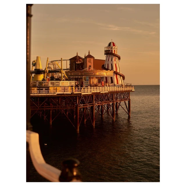 A long pier extending over the ocean, topped with amusements and a lighthouse-like structure in red and white, at sunset.