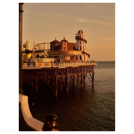 A long pier extending over the ocean, topped with amusements and a lighthouse-like structure in red and white, at sunset.