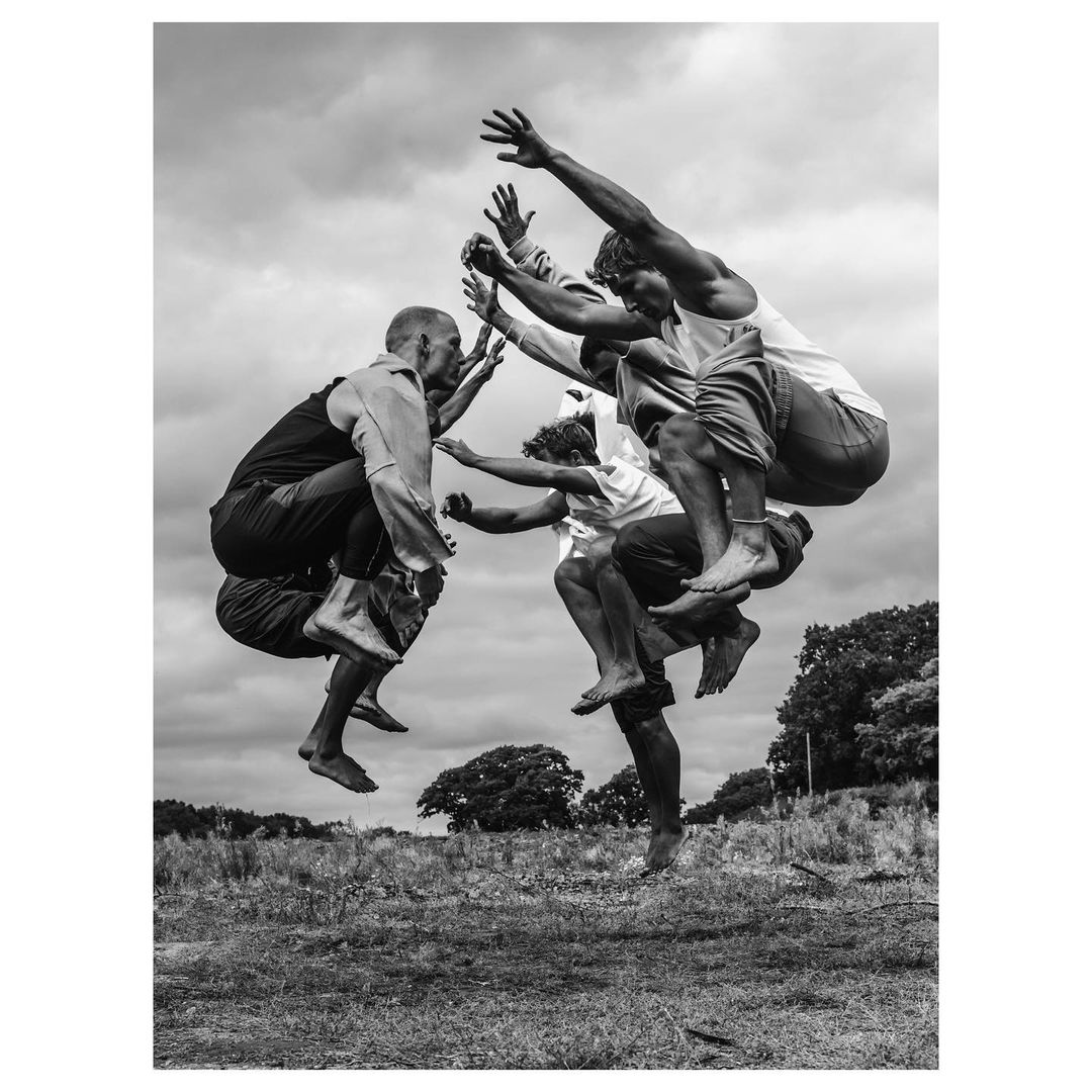 A black and white shot capturing five adult men mid-air, seemingly jumping over one another in a grassy field under a cloudy sky.