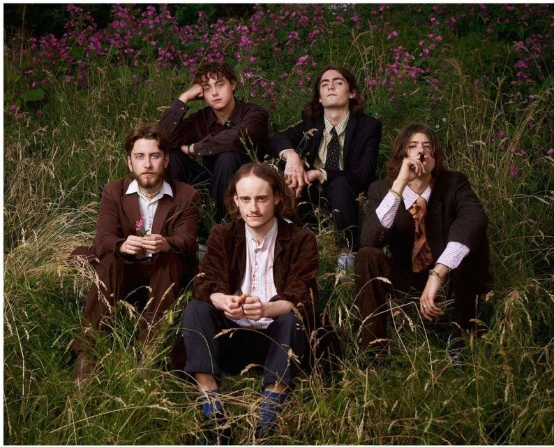 Five adult males sit in tall grass in a field with flowering plants, all wearing suits, in an outdoor setting.