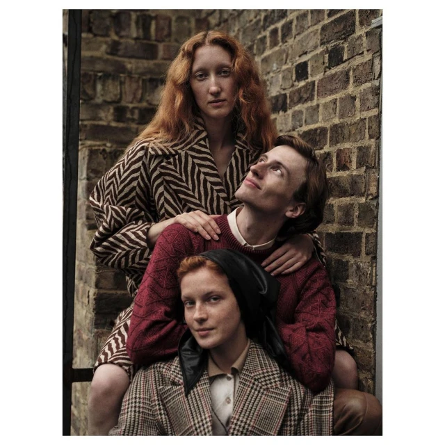 Fashion shot of three models outdoors. A woman with red hair is seated, with a man and another woman in front of a brick wall backdrop.