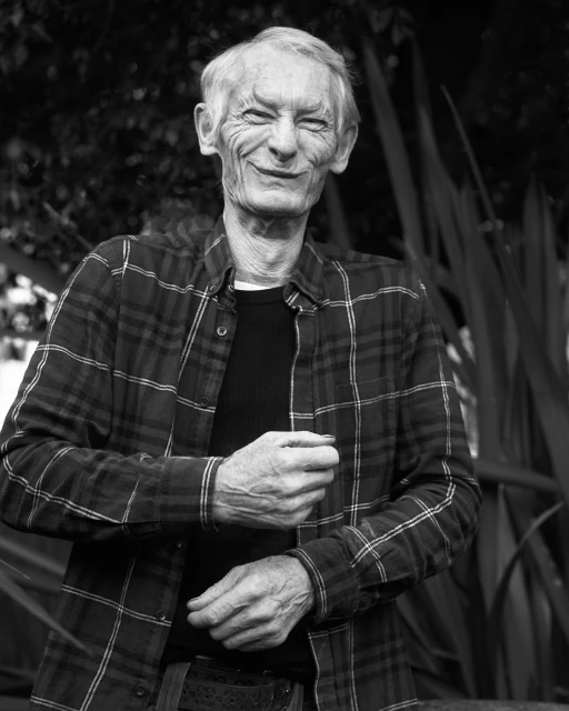 Black and white portrait of an elderly man smiling with wrinkles, wearing a flannel shirt, in an outdoor environment with foliage.