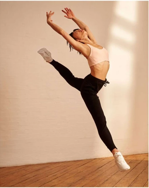 A woman in a pink top and black pants leaps with her arms overhead in a dance pose in a studio with a wood floor and white walls.