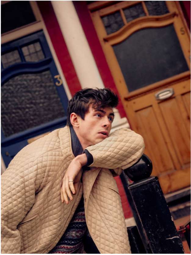 Stylish young man in a quilted jacket leans against a railing outdoors with colorful buildings in the blurred background.