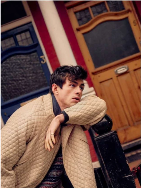 Stylish young man in a quilted jacket leans against a railing outdoors with colorful buildings in the blurred background. 