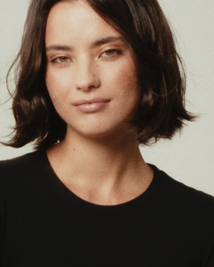 Portrait of an adult woman with short dark hair and a black top, posing in front of a neutral background in a close-up shot.