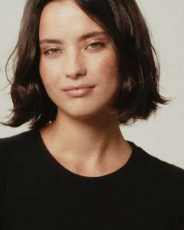 Portrait of an adult woman with short dark hair and a black top, posing in front of a neutral background in a close-up shot.