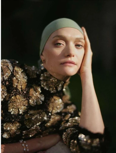 Close-up portrait of a fair-skinned adult female with a head covering, wearing a sequined top, posing outdoors with a blurred background.