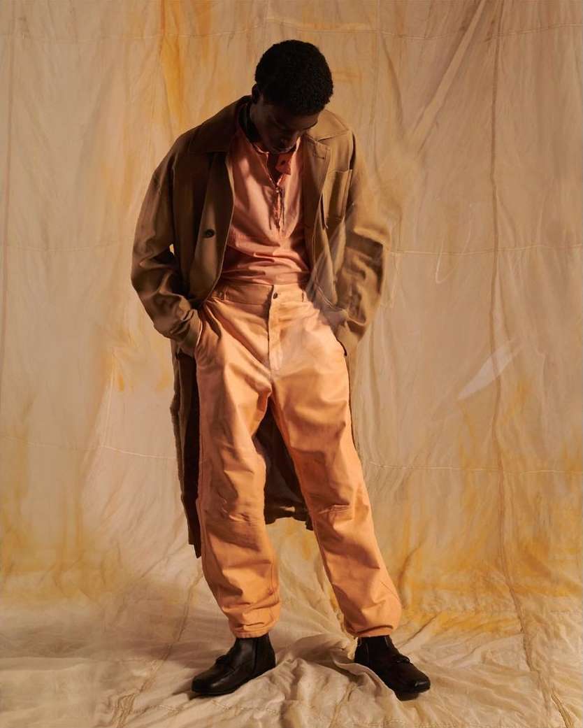 Dark-skinned male model in pink shirt and pants with a tan trench coat poses in a studio with a mottled tan background.
