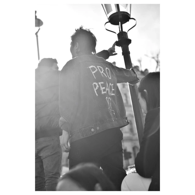 Back view of a person in a denim jacket with 'Pro Peace' written on it, standing next to a lamp post with other people nearby.
