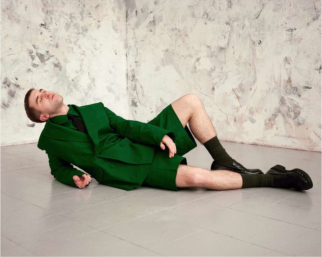 Man in green suit with shorts lies on his side on a gray floor against a textured wall. He wears black socks and shoes.
