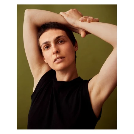 Close-up portrait of a woman with short dark hair in a black sleeveless top with arms raised, against a dark olive background.