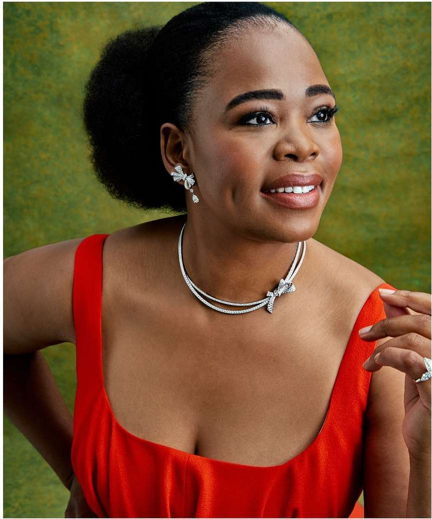 Close-up of a dark-skinned woman in an orange dress with diamond jewelry smiling against an abstract green background, portrait orientation.