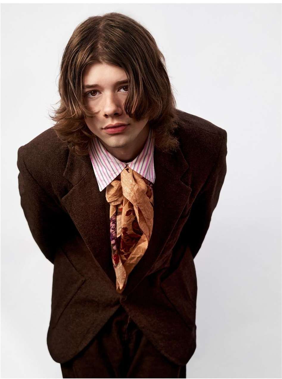 Portrait of a young adult male with brown hair, wearing a brown suit jacket and patterned neck scarf, against a white background.