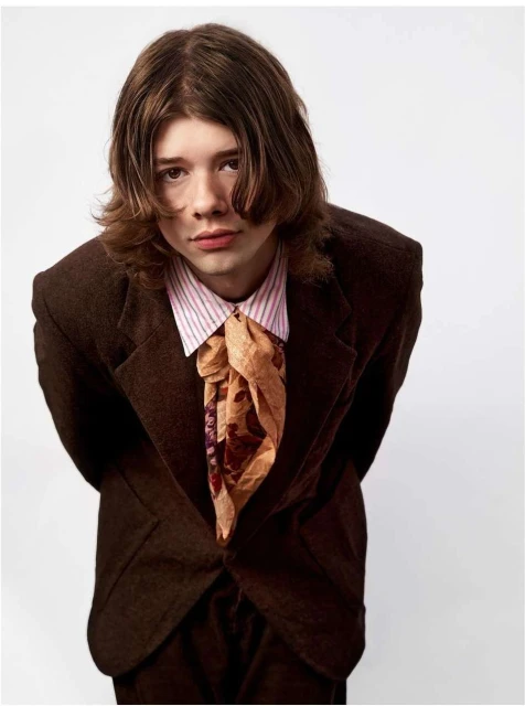 Portrait of a young adult male with brown hair, wearing a brown suit jacket and patterned neck scarf, against a white background.