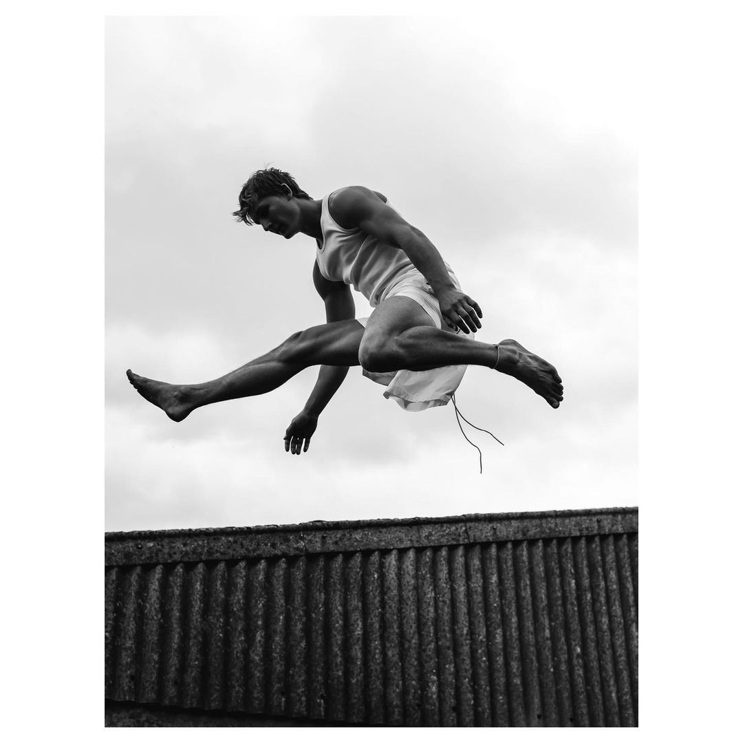 Muscular man in mid-air leap on corrugated metal roof with cloudy sky. Black and white athletic fashion shot for fitness or sport.