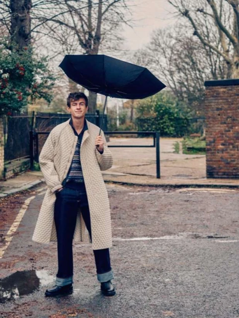 A young adult man stands on a wet asphalt road holding an umbrella, wearing a long patterned coat, jeans, and black shoes, outdoors.