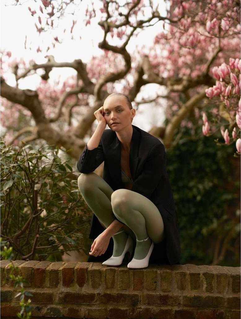 Woman in blazer crouching against brick wall in outdoor setting with flowering tree behind; the lighting is soft and diffused.