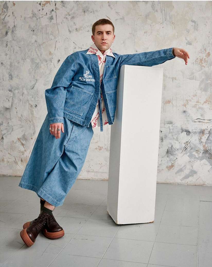 Male fashion model in denim jacket and wide leg jeans posing in studio against a textured wall next to a white column.