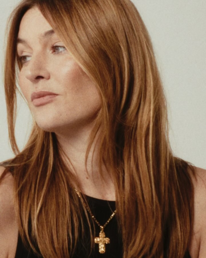 Close-up portrait of a woman with long auburn hair wearing a black top and gold cross necklace against a neutral background.