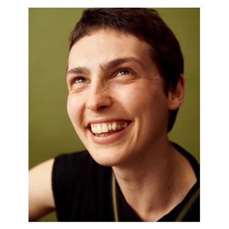 Portrait of a smiling adult woman with short hair. She is wearing a black sleeveless shirt against an olive green background.