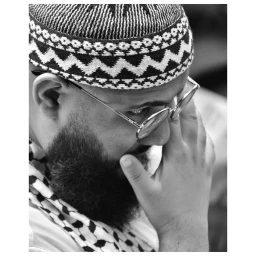 Close-up portrait of a man wearing round glasses, a patterned kufi cap, and a keffiyeh scarf by Virginie Khateeb