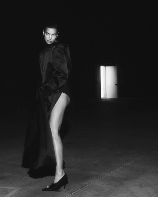 Black and white studio portrait of a woman in a jacket dress and heels against a dark backdrop and light from a doorway.