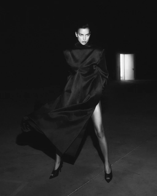 Black and white portrait of a woman in a draped dress and heels stands in front of a door in a dark room, fashion editorial.