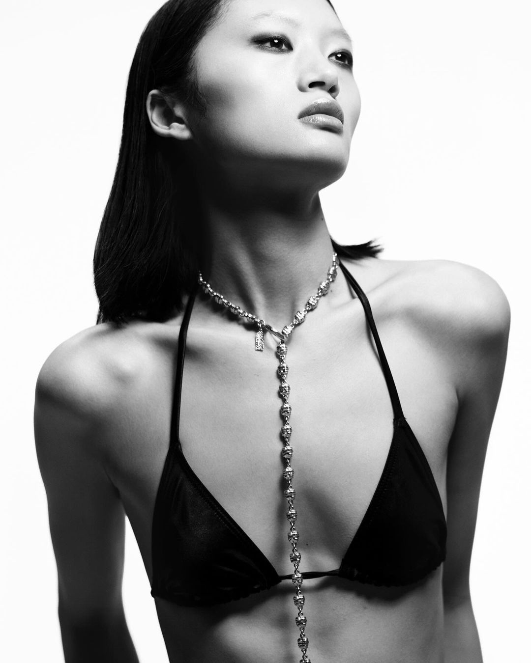 Monochrome portrait of an Asian woman in a bikini top and necklace, against a bright white background, looking up and to the right.